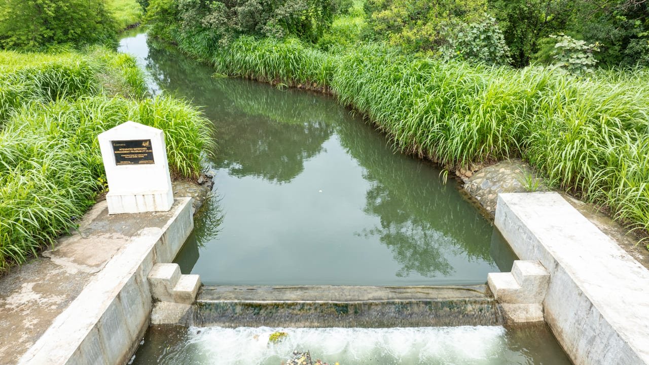 A concrete check dam with water spilling over its weir, a project plaque on the bank