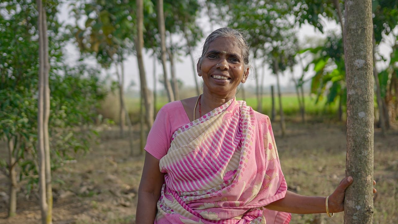 An elderly woman in a pink saree smiling as she leans against a tree in an orchard