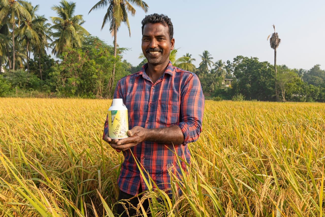 A smiling farmer holding a bottle of bio-input in a golden paddy field
