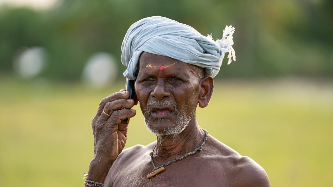 An elderly farmer in a white turban talking on a mobile phone beside his field