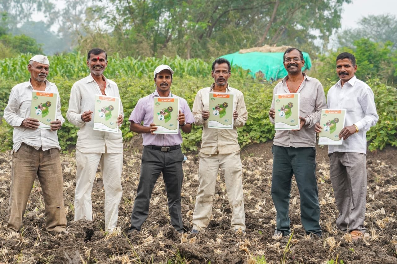 Six farmers in a ploughed field holding up printed soil health cards