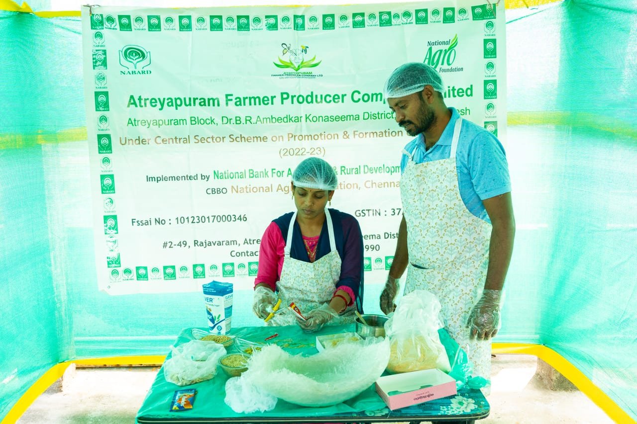 Two FPO workers in hairnets packing food products in front of their FPO's banner
