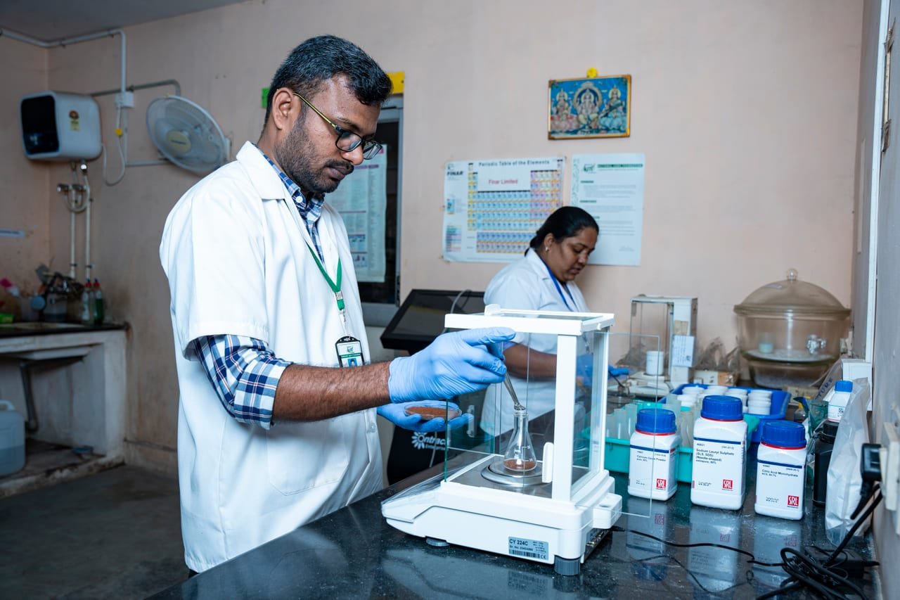 A laboratory technician weighing a sample on an analytical balance, a colleague behind
