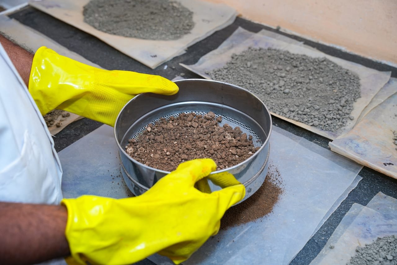 Gloved hands holding a steel sieve of soil over trays of processed samples