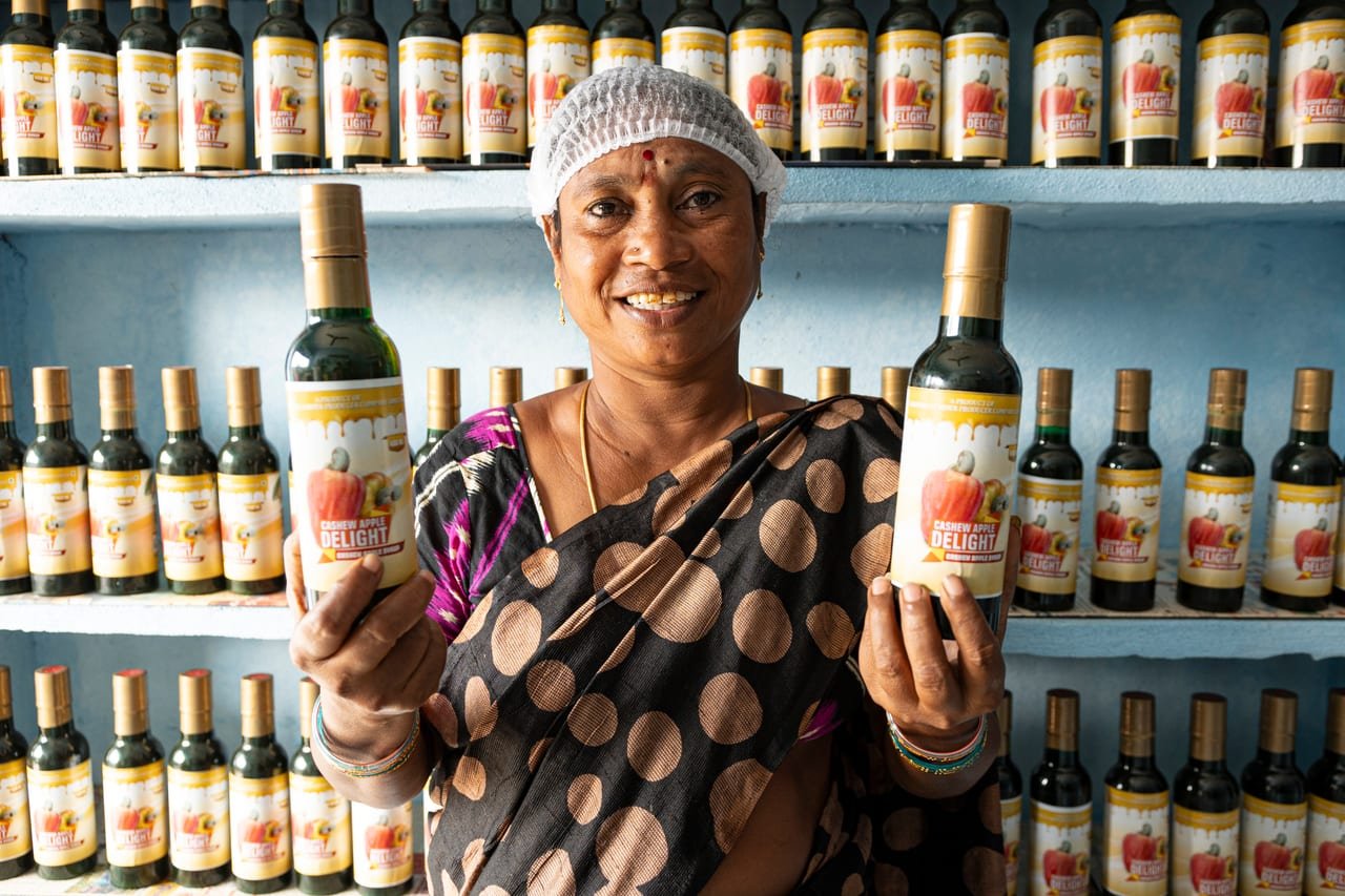A smiling woman in a hairnet holding bottled products in front of stocked shelves