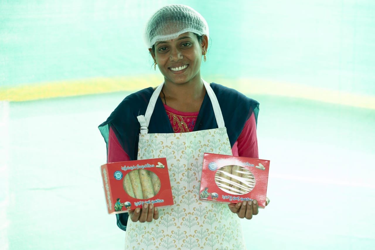 A smiling woman in a hairnet and apron holding packaged food products