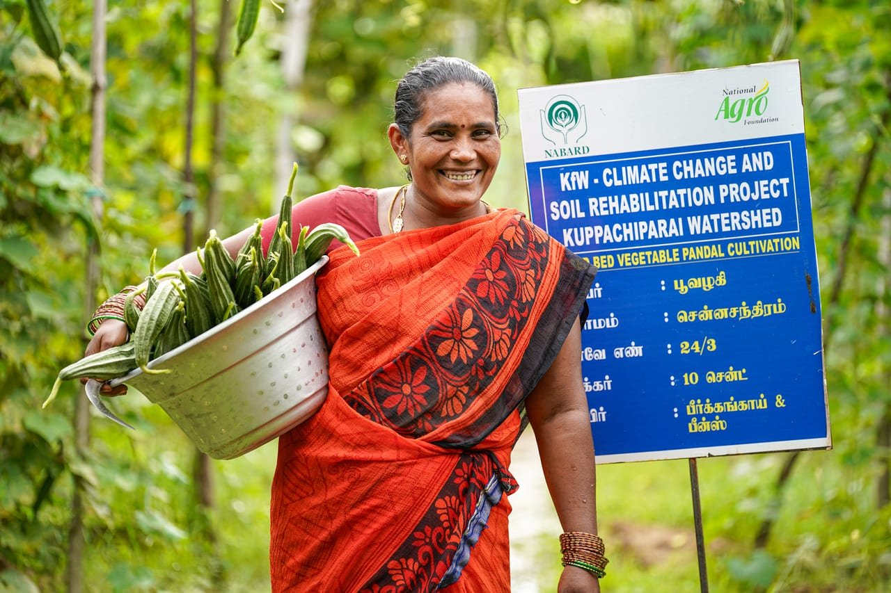 A smiling woman carrying a basin of freshly harvested ridge gourds beside a project board