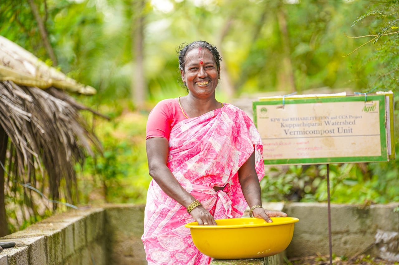 A smiling woman holding a yellow tub beside a vermicompost unit and its project board