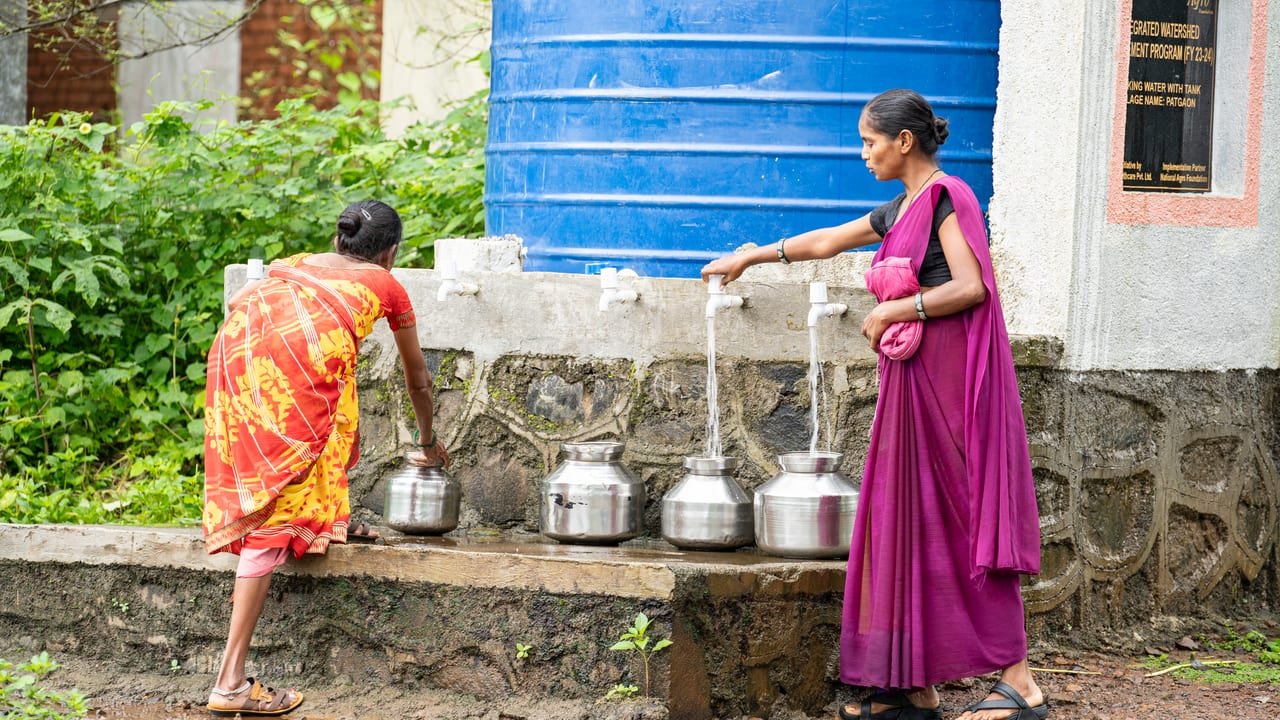 Two women filling steel pots from the taps of a community drinking-water tank