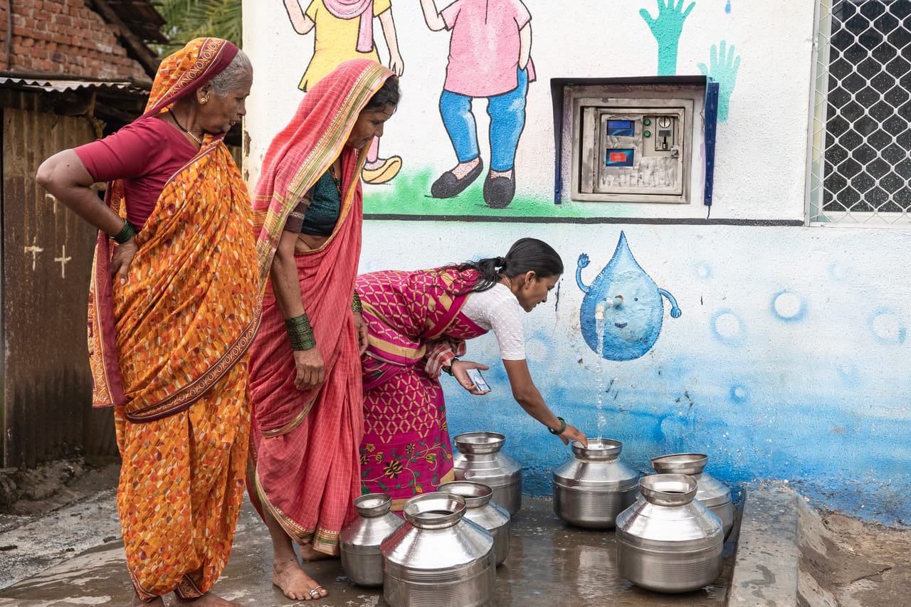 Women filling steel water pots at a village drinking-water kiosk