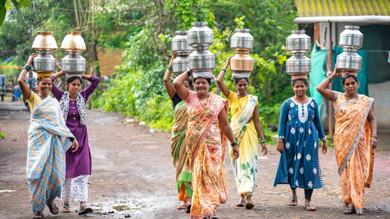 Women walking along a village road with stacked steel water pots on their heads
